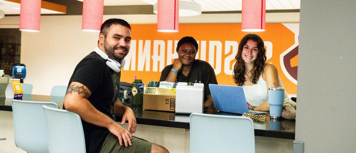 Image of three students sitting at a table in Hawk Nest smiling and looking at the camera.